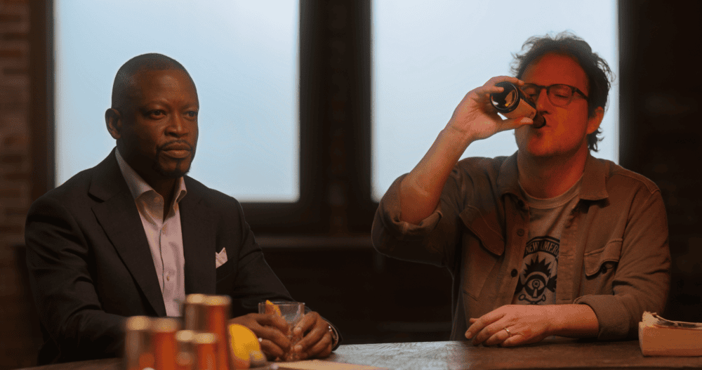 Two men sitting at a bar with one drinking already and the other just sitting.
