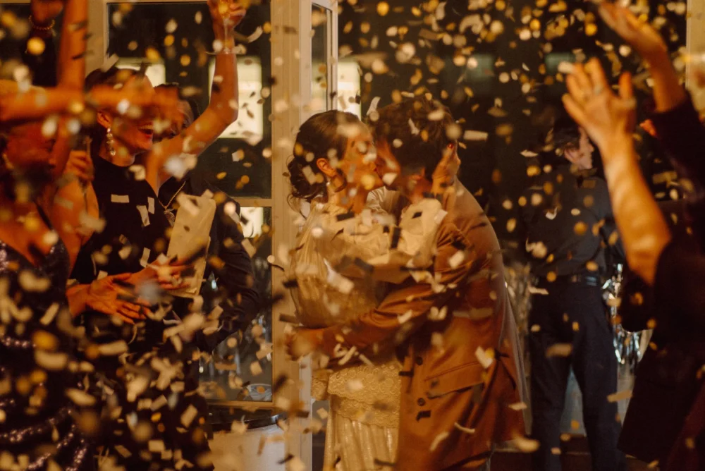 A bride and groom kissing at a wedding with flowers being thrown at them by friends.